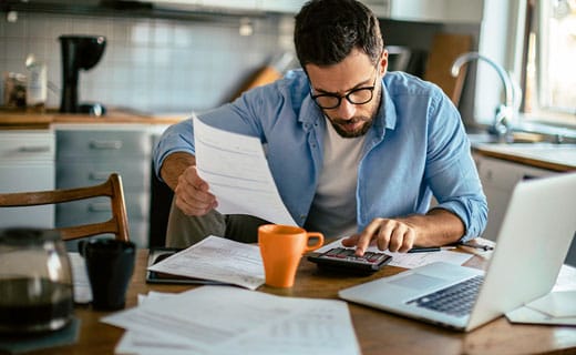 Man sitting at kitchen table surrounded by papers and laptop, using a calculator.